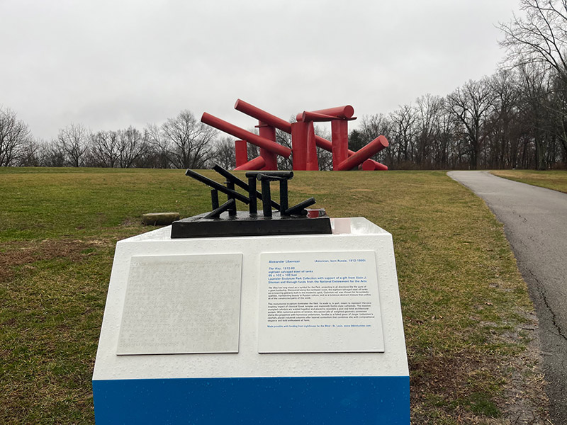 A large sculpture with various red cylinders stacked around and on each other on a barren field. In front is a miniature of that sculpture on a podium.