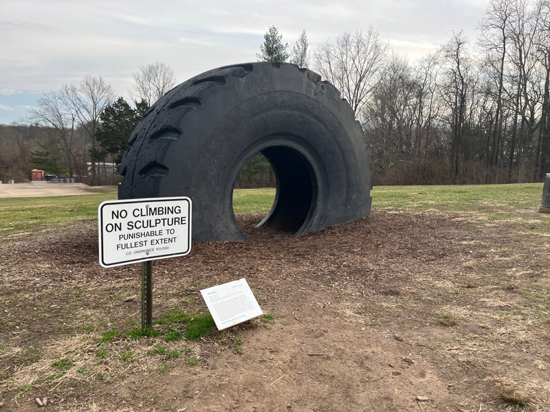 A comically huge tire sits partially buried in mulch on a patchy field. A sign nearby says, 'No climbing on sculpture. Punishable to fullest extent.'