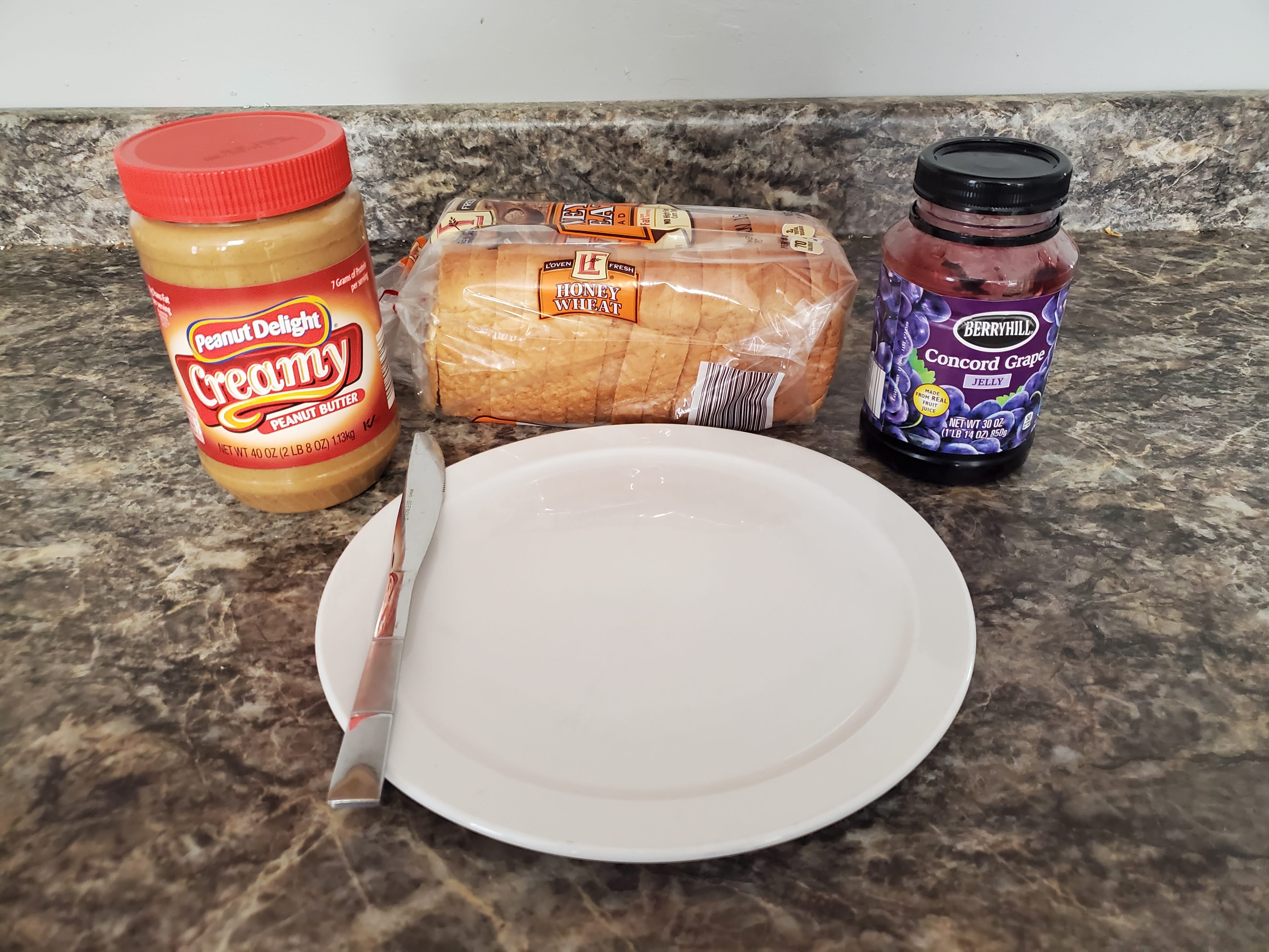 On a countertop lies peanut butter, a loaf of bread, jelly, and a plate with a butter knife on top.