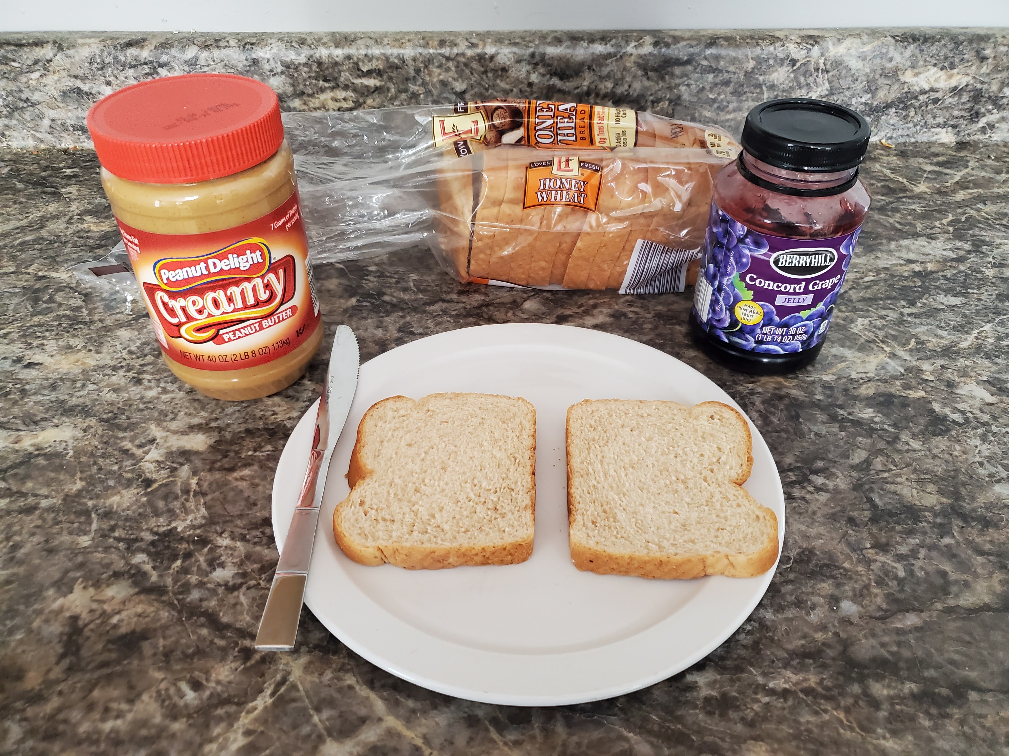 On a countertop lies peanut butter, an opened loaf of bread, jelly, and a plate with two slices of bread and a butter knife on top of it.