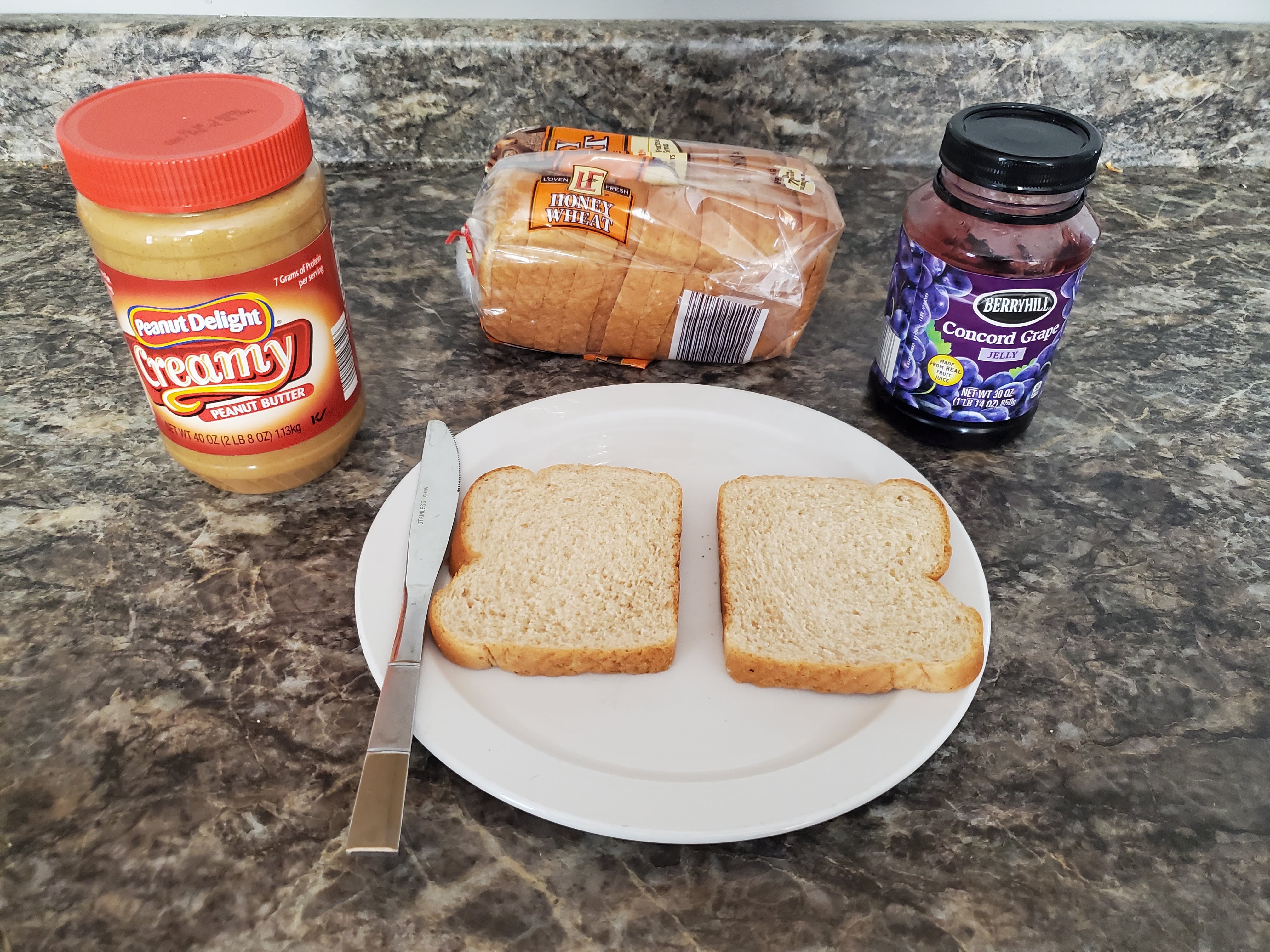 On a countertop lies peanut butter, a closed loaf of bread, jelly, and a plate with two slices of bread and a butter knife on top of it.