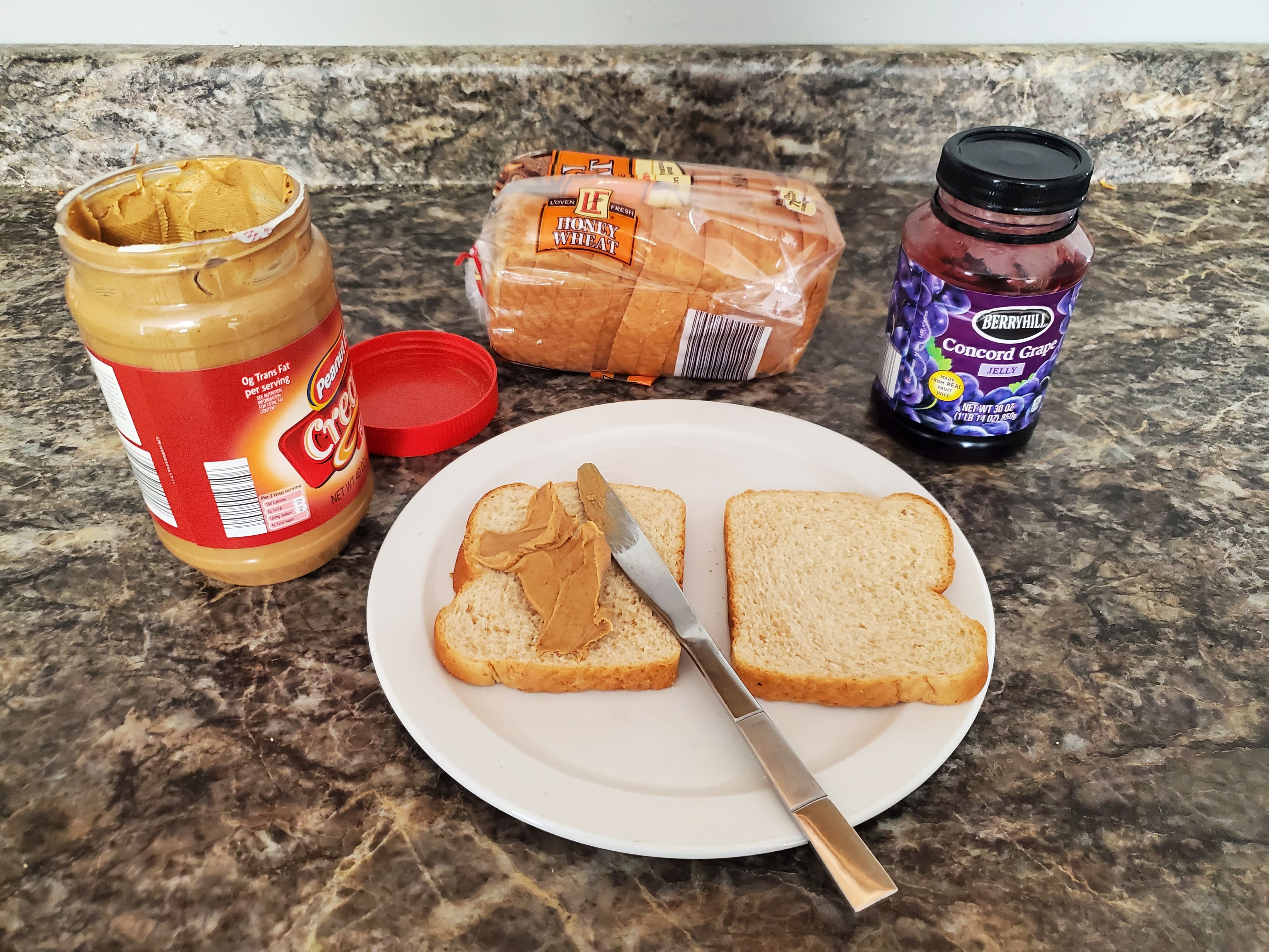 On a countertop lies an opened jar of peanut butter, a loaf of bread, jelly, and a plate with two slices of bread and a butter knife on top of it. The slice on the left has a dollop of peanut butter on it.