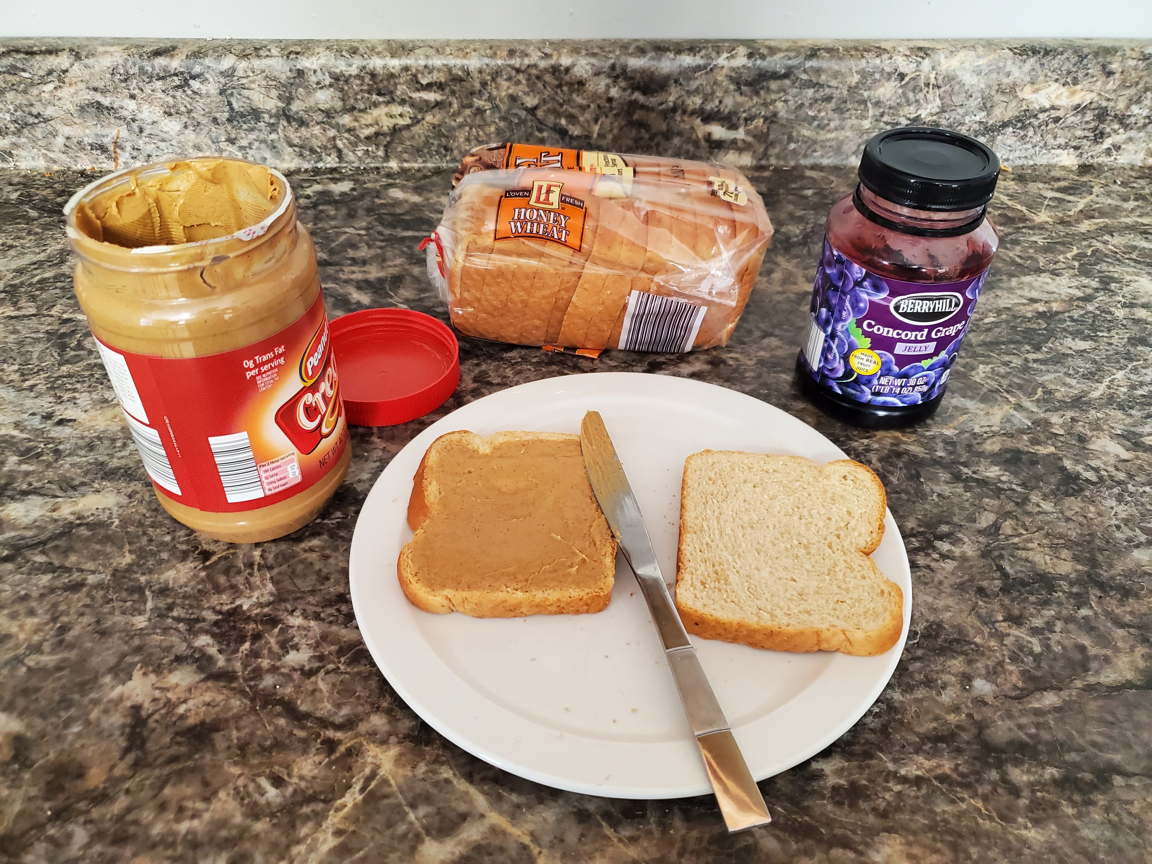 On a countertop lies an opened jar of peanut butter, a loaf of bread, jelly, and a plate with two slices of bread and a butter knife on top of it. The slice on the left has had peanut butter spread across it.