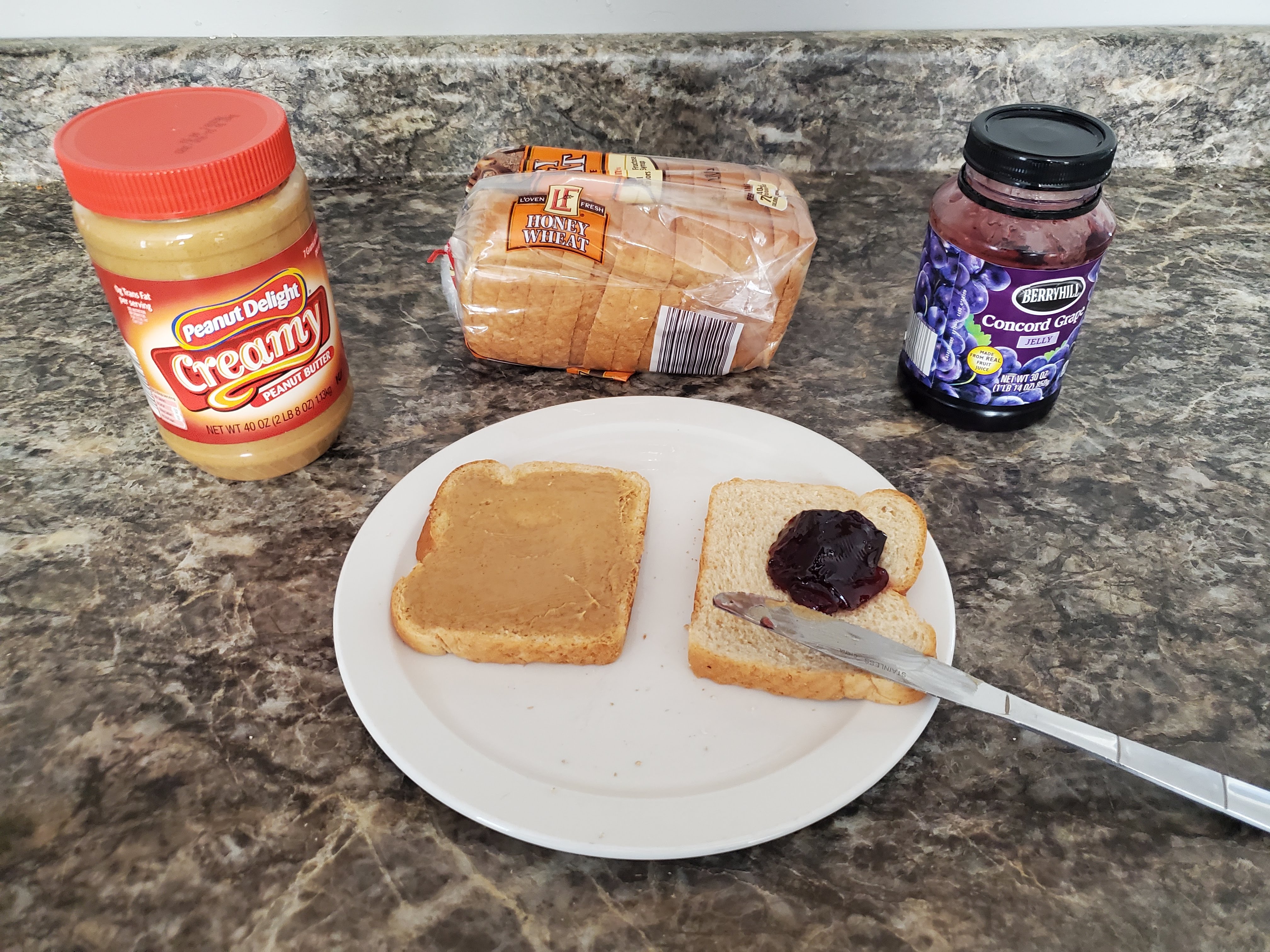 On a countertop lies a of peanut butter, a loaf of bread, an opened jar of jelly, and a plate with two slices of bread and a butter knife on top of it. The slice on the right has a dollop of jelly on it.