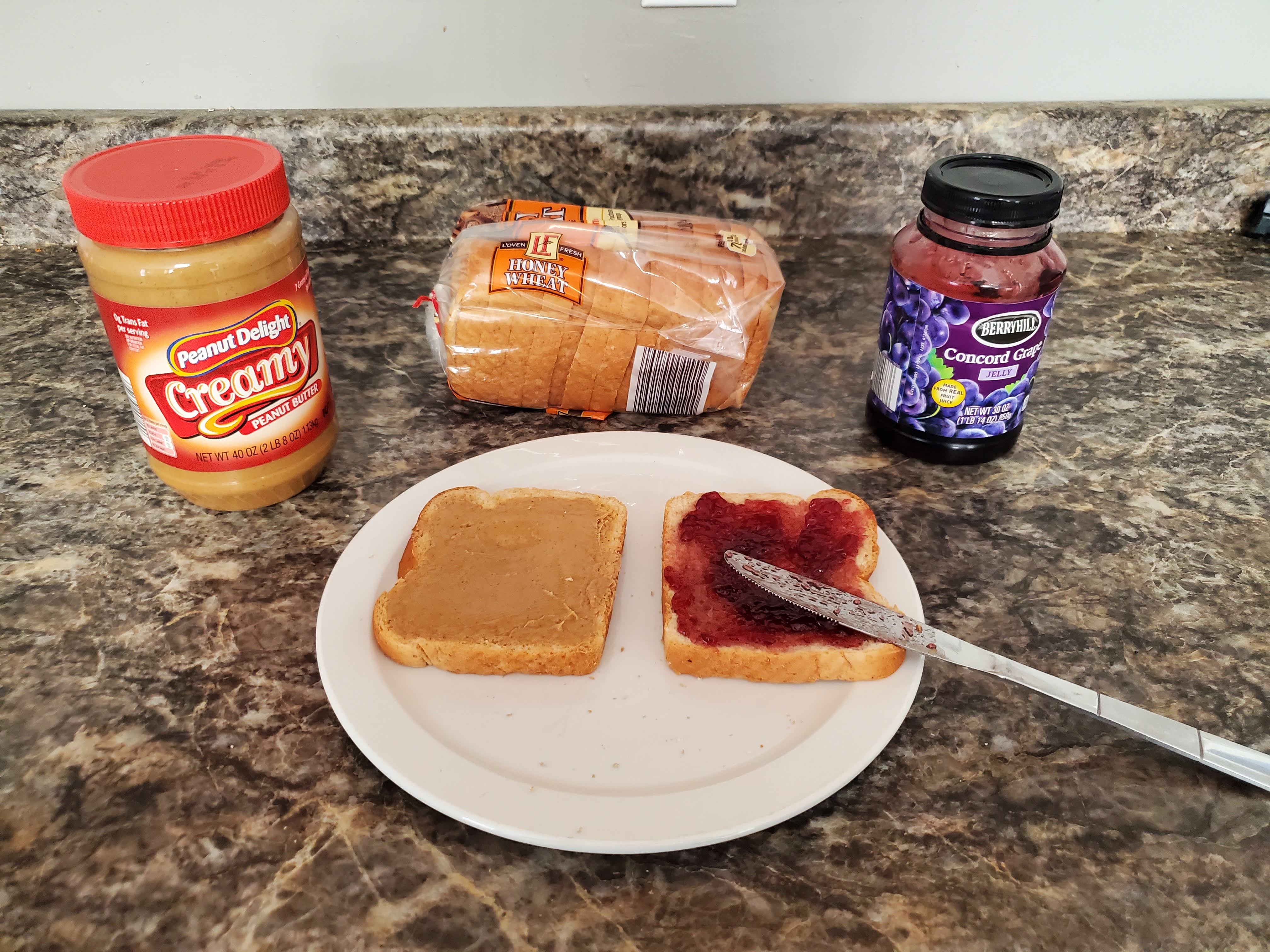 On a countertop lies an opened jar of peanut butter, a loaf of bread, jelly, and a plate with two slices of bread and a butter knife on top of it. The slice on the right has had jelly spread across it.