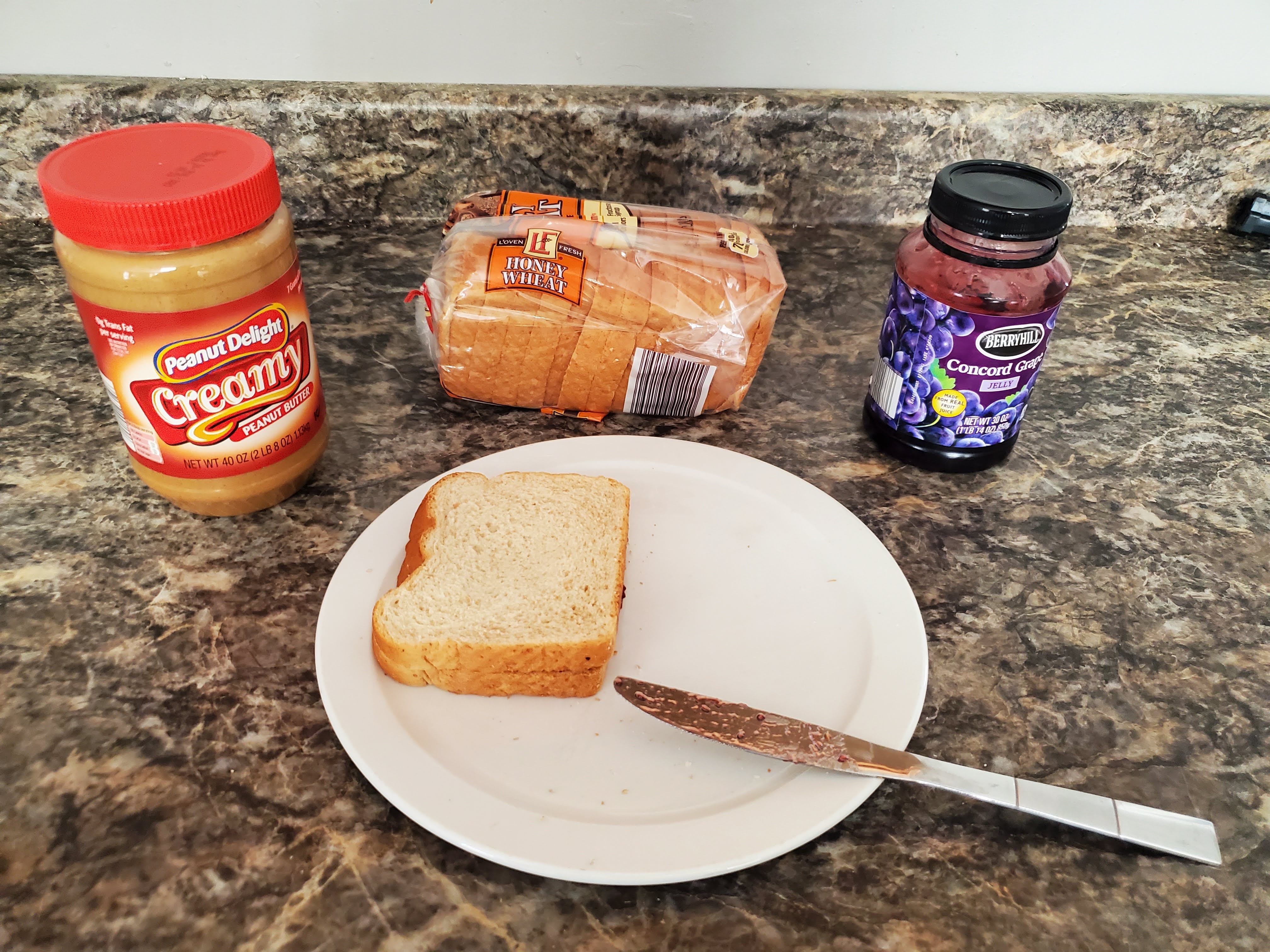 On a countertop lies an opened jar of peanut butter, a loaf of bread and jelly behind a plate. On the plate, two slices of bread have been put together to create a peanut butter and jelly sandwich.