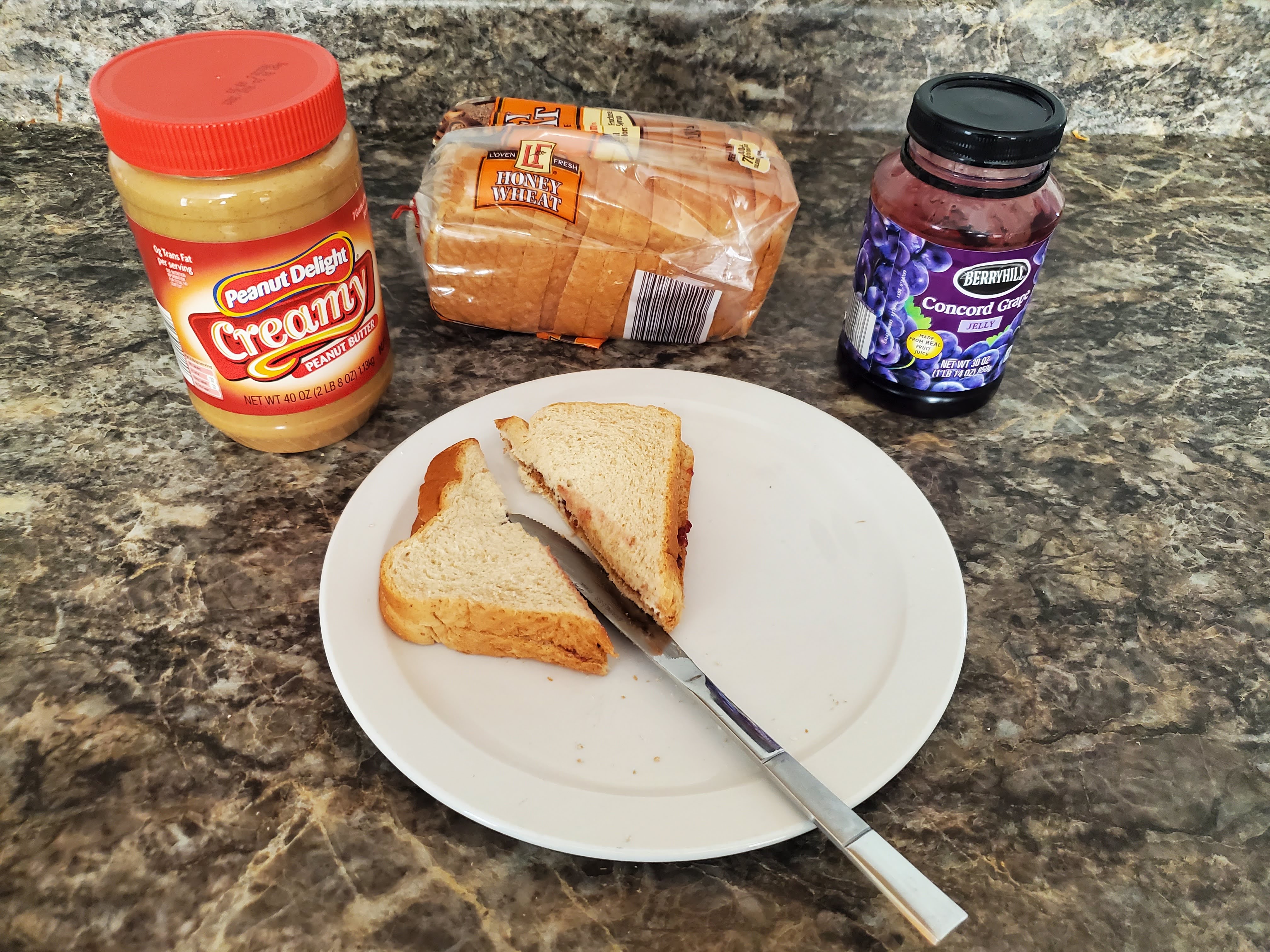 On a countertop lies an opened jar of peanut butter, a loaf of bread and jelly behind a plate. On the plate, two slices of bread have been put together and sliced in half with a butter knife to create a peanut butter and jelly sandwich.